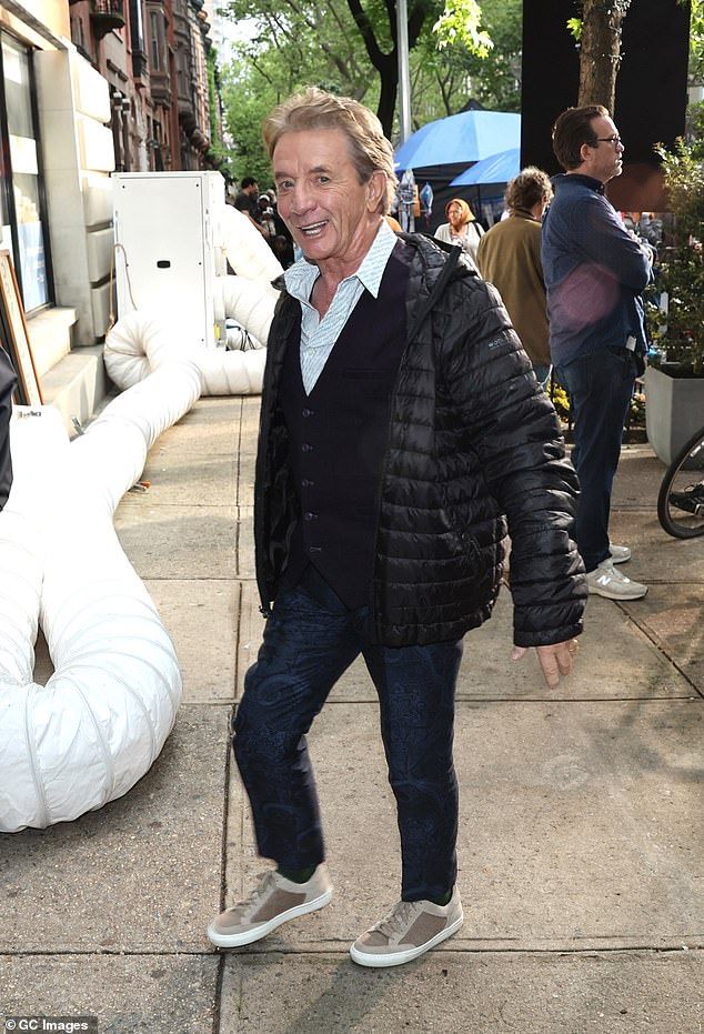 Meanwhile, Martin bundled up in a padded black coat, a black button-up cardigan, and a striped shirt, smiling for the cameras during a quick break from filming